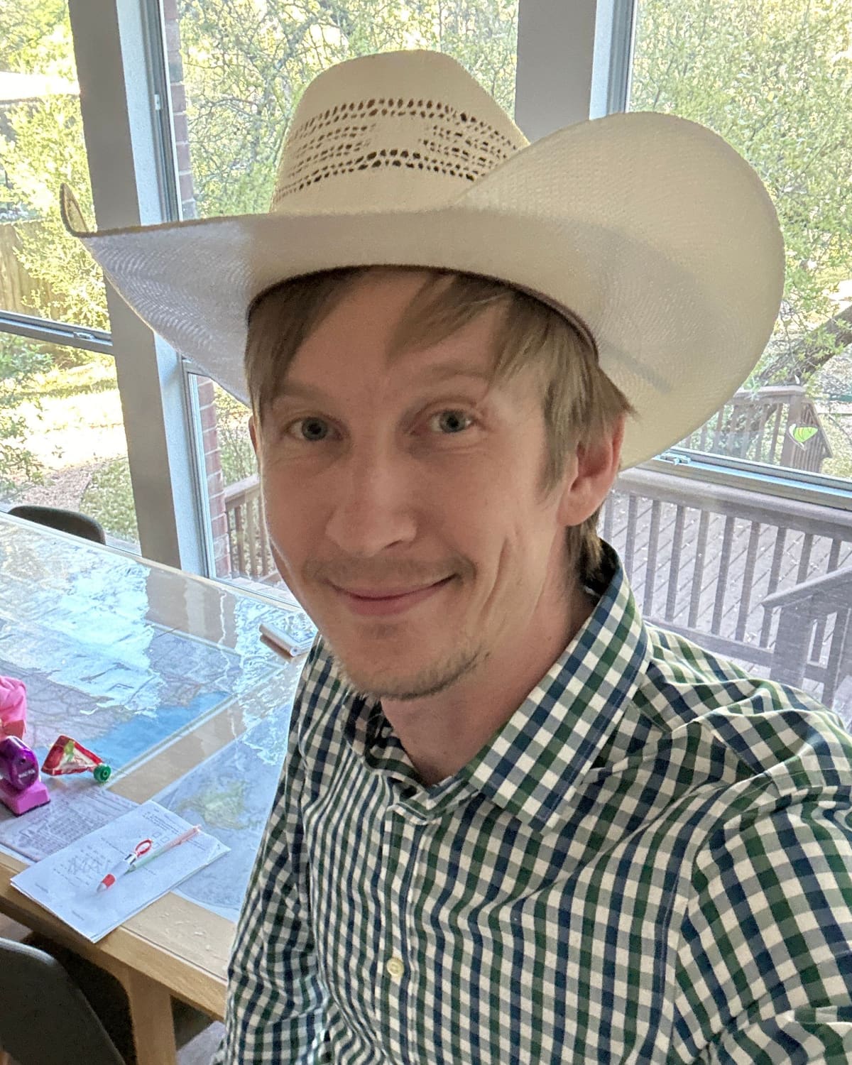 Misha smiling in a white straw cowboy hat and a green-and-blue gingham shirt, at home by windows onto a leafy backyard